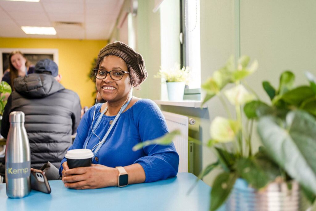 Adult student wearing a brown hat and blue jumper, sat with a coffee smiling