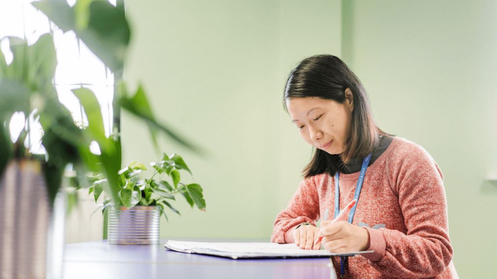 Adult female learner sat at a table with plants on it working and writing in her text book