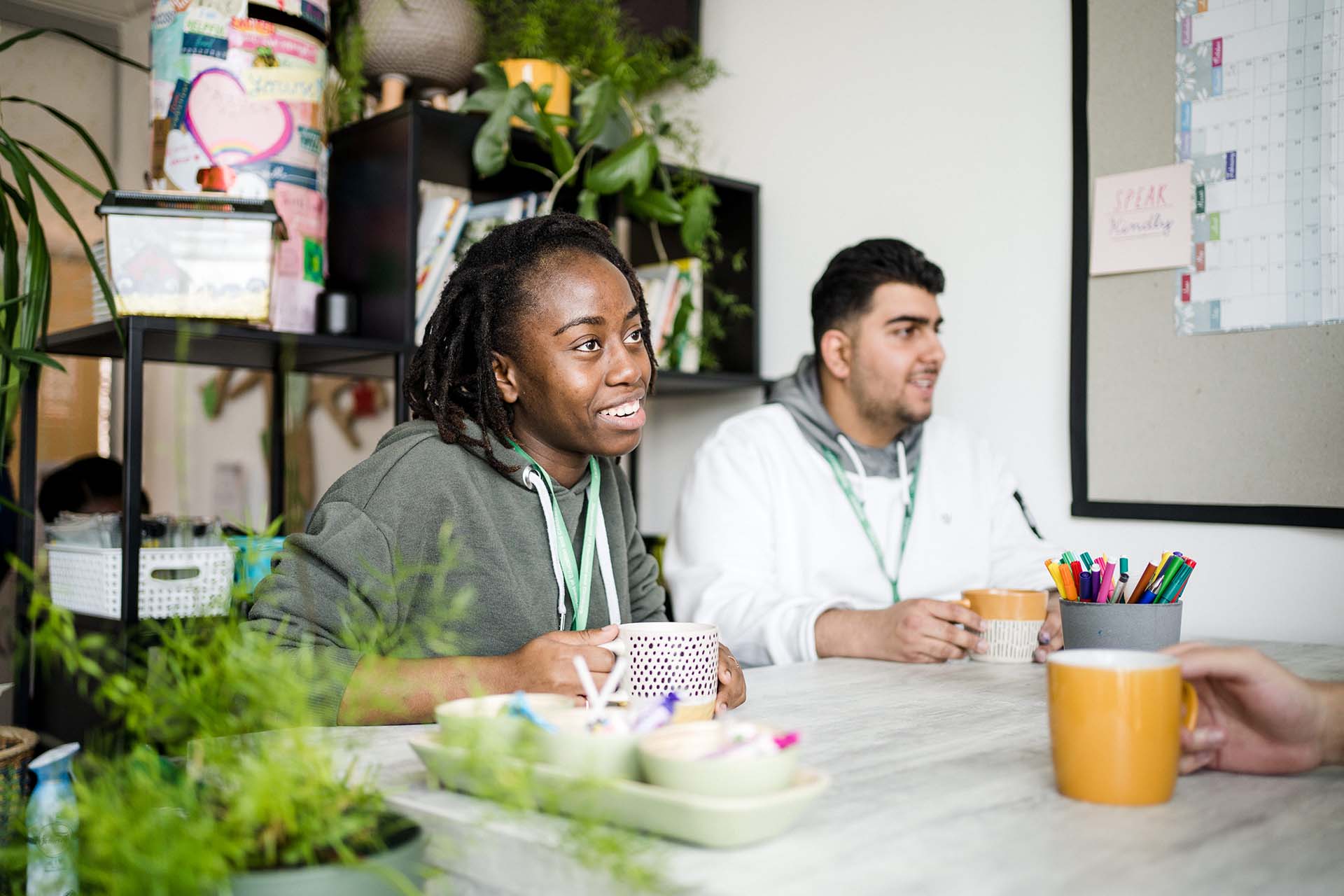 Two students wearing lanyards and smiling, sat having a cup of tea at a table in a room with plants, books and pens.