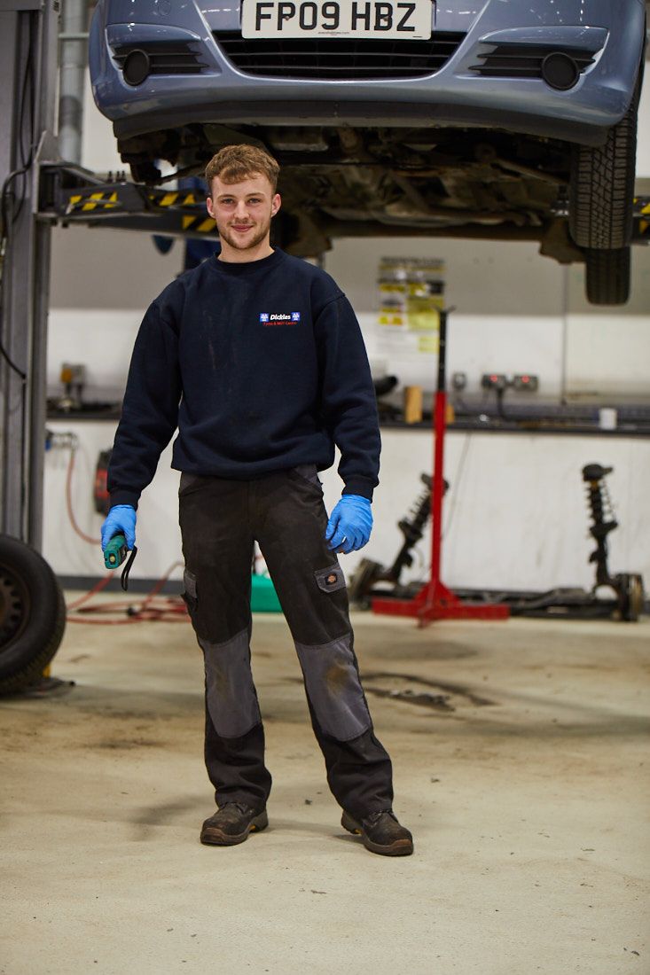 LCC engineering student on an automotive course looking at the camera with a car on a lift behind raised to be able to work under