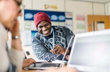 Adult student wearing a lanyard and beanie hat sat next to someone else in a classroom smiling at a laptop