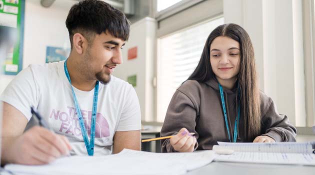 Two students wearing Leeds Sixth Form College lanyards sat at a classroom desk looking at worksheets together