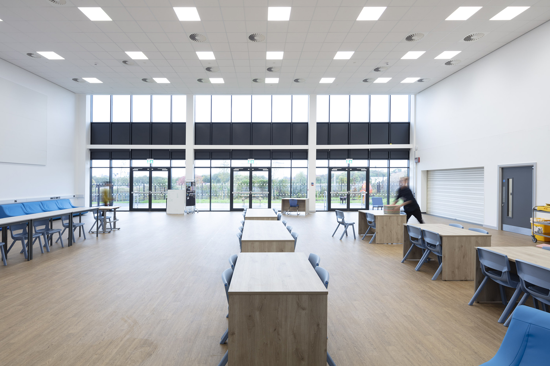 Open-plan canteen with desks and chairs and three double glass doors leading to outside