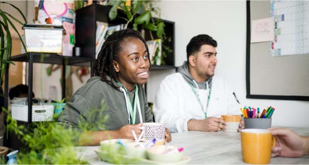 Two Leeds City College students sat smiling wearing lanyards, having a cup of tea in a classroom with plants