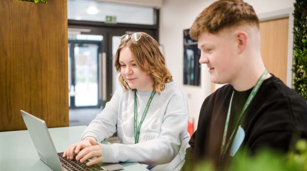 Two students wearing Leeds City College lanyards sat down looking at a laptop together and smiling
