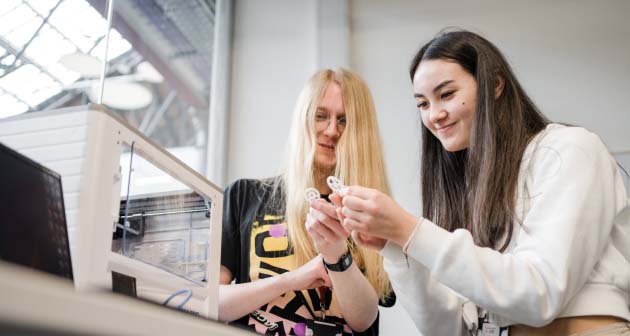 Two students with long hair smiling looking down at 3d printer cogs