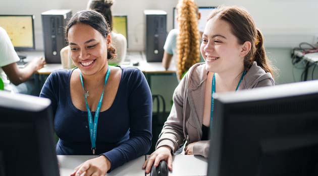 Two people wearing Leeds City College lanyards sat in a computer room looking at a computer screen smiling