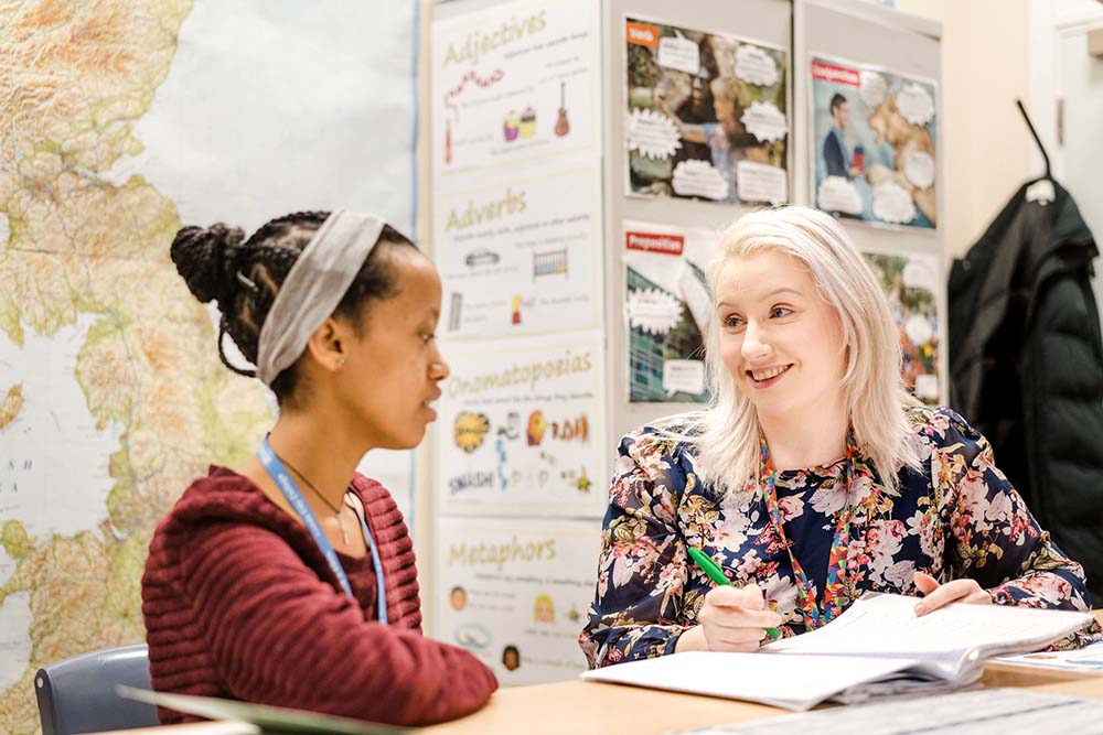 Teacher sat with adult student in a classroom smiling whilst discussing a course book