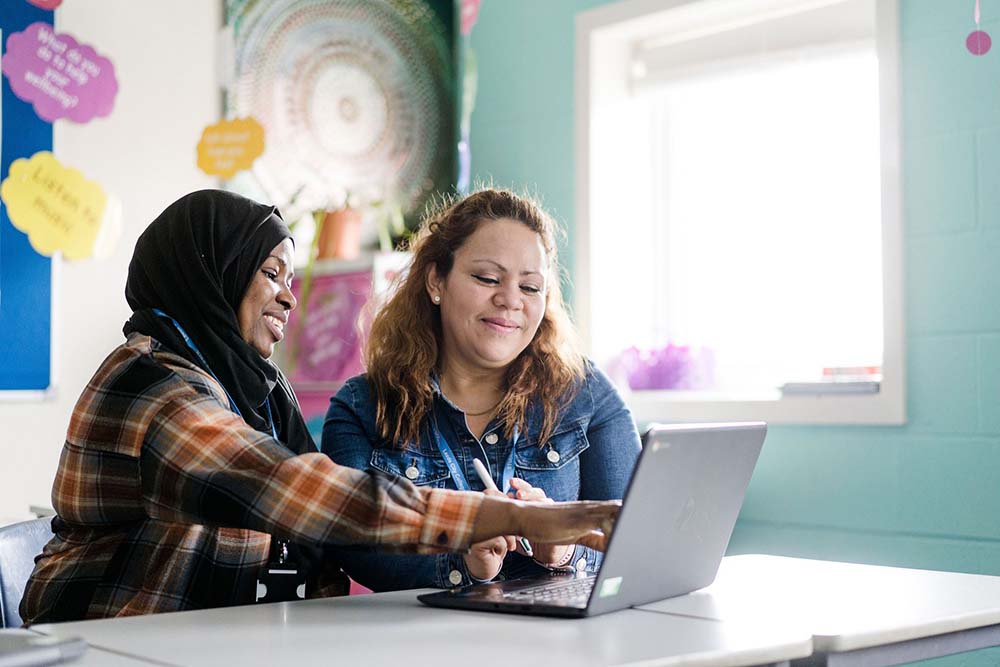 Teacher and adult student sat in a bright classroom looking at a laptop screen together and smiling