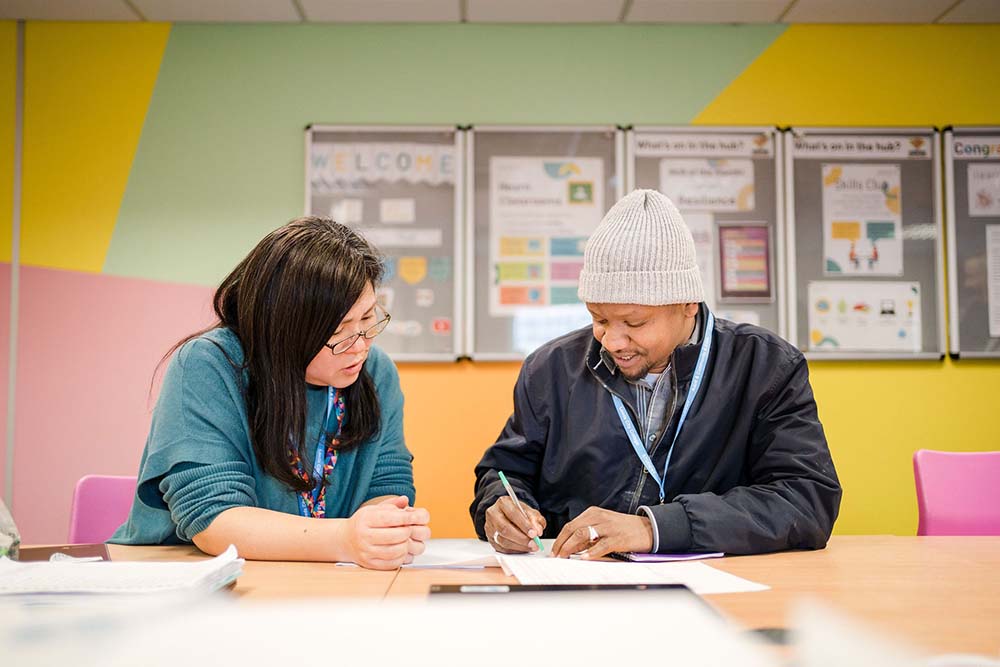 Teacher sat with adult student together looking down in discussion whilst the student is writing in a course book