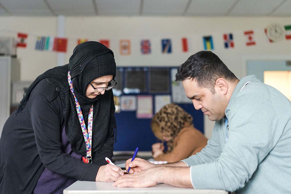 Teacher helping adult student as he writes in a course book sat at a desk in a classroom, both looking down with pens concentrating