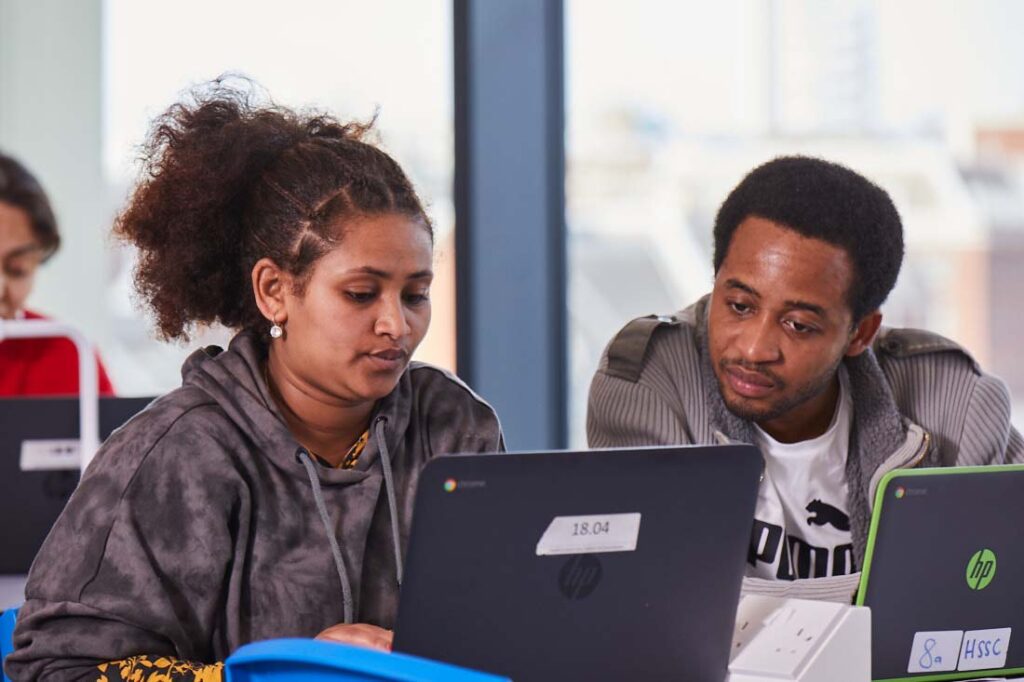 Two adult students sat in a classroom in front of two laptops looking at a screen together