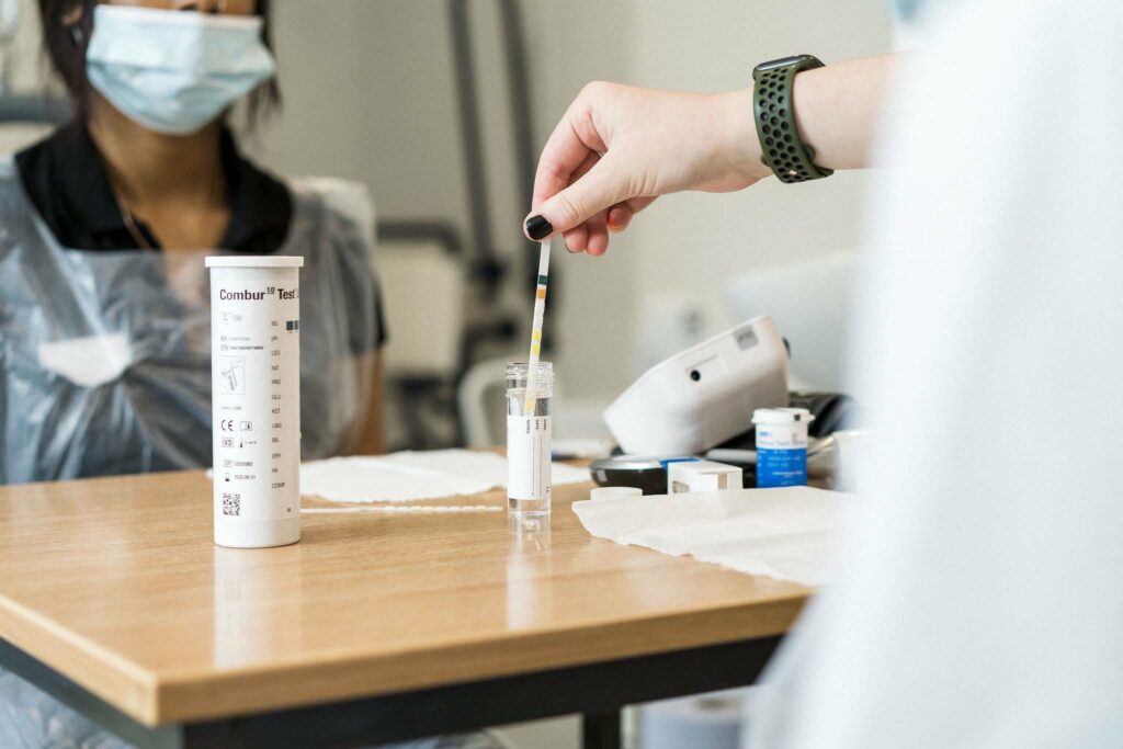 A hand is using pipette to squeeze liquid out of a test tube in a medical science room. A person is sat behind wearing a face mask and plastic overalls.