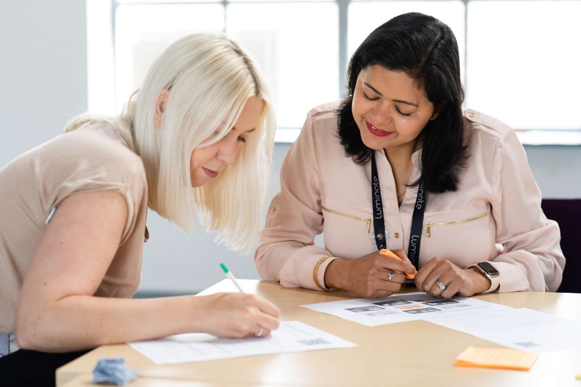 A staff member with medium length black hair wearing a pink jacket assisting a woman with medium length blonde hair and a light beige top with a form