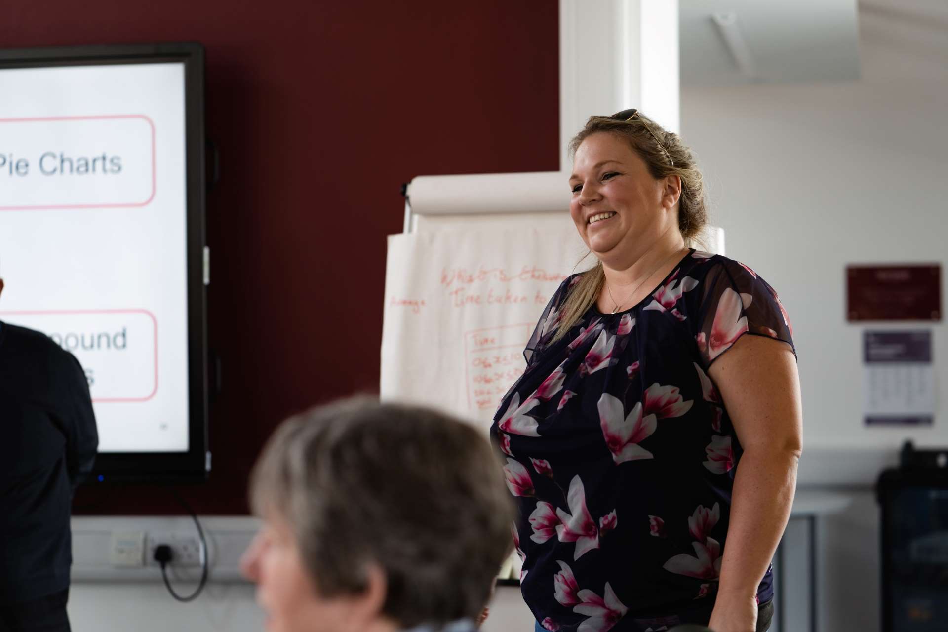 A woman stood at the front of the class room delivering a presentation wearing a floral pattern black top and shades pushed up into her hair