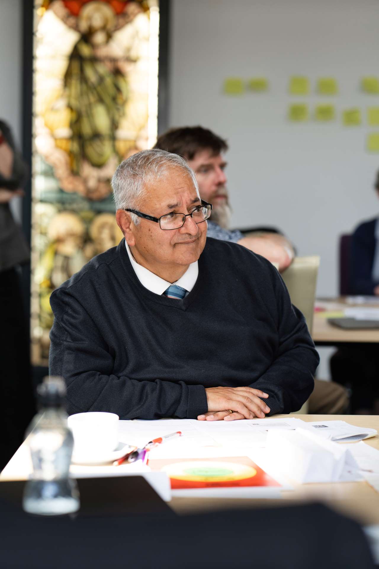 A older adult learner sat down at a desk looking to his right he has white thinning hair and glasses with a cardigan jumper with tie and shirt