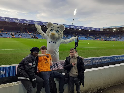 Three students sat by a football pitch and with a team mascot