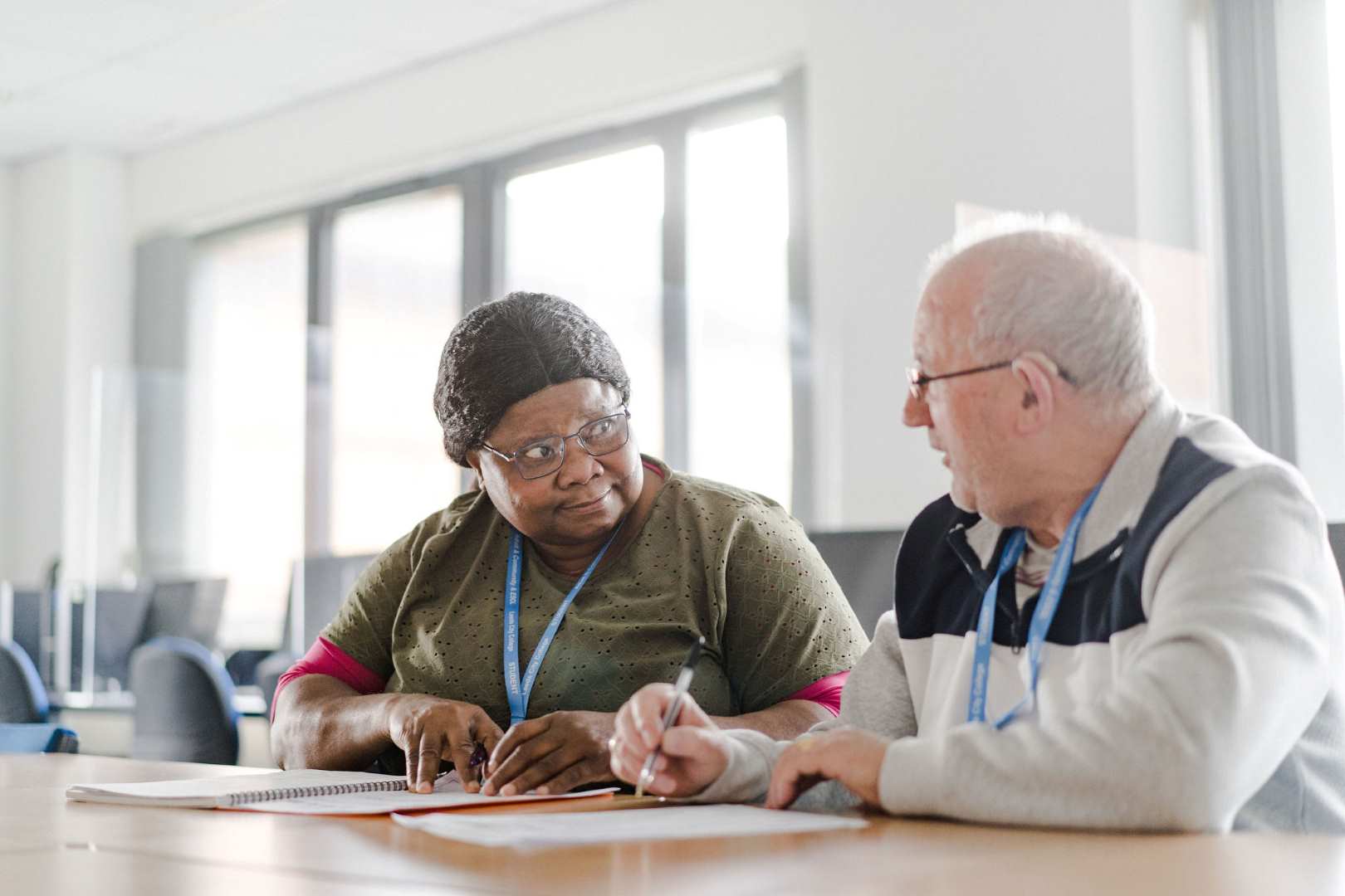 Two older adult learners both holding pens sat at a desk, one with a note book for notes and another with a printed document. Looking at one another discussing what they are doing