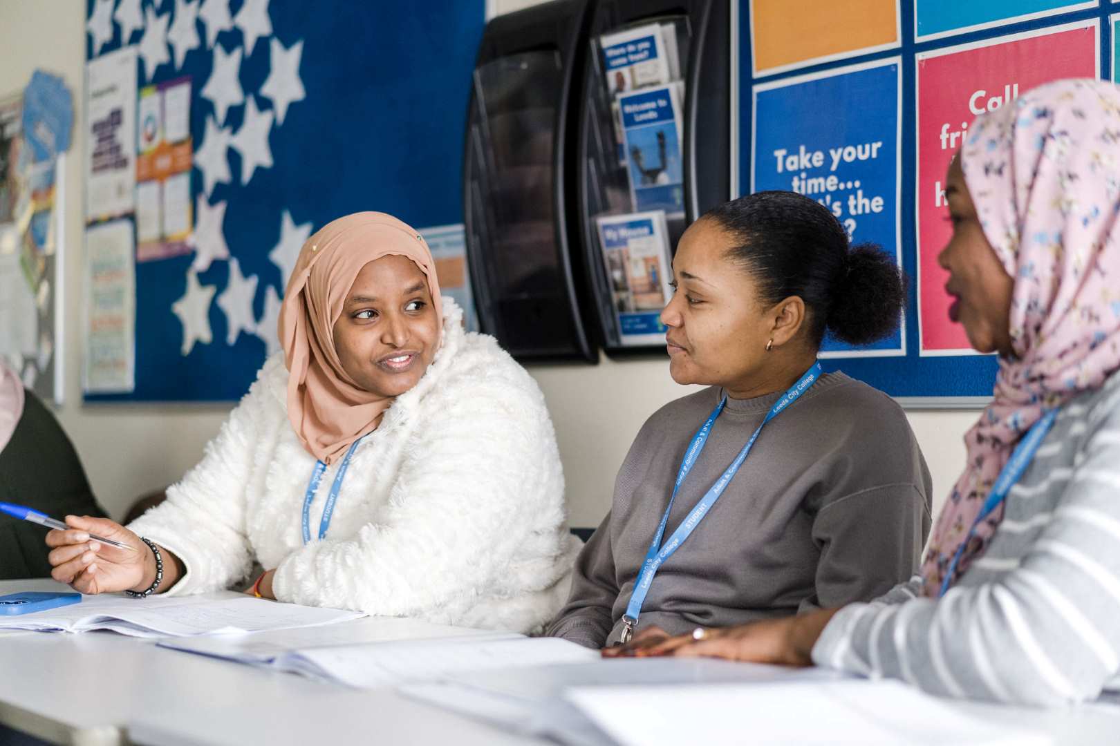 Three adult learners sat in a class together with text books as they are happily discussing their work