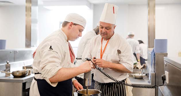 Catering student with Head Chef learning to prepare food in the Printworks kitchen, both wearing kitchen uniforms and chef hats