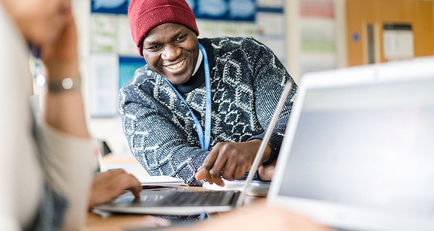 Adult student wearing a lanyard and beanie hat sat next to someone else in a classroom smiling at a laptop