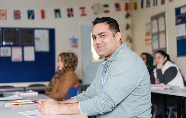 Adult learner sat in classroom smiling and looking at the camera with learners behind sat down out of focus