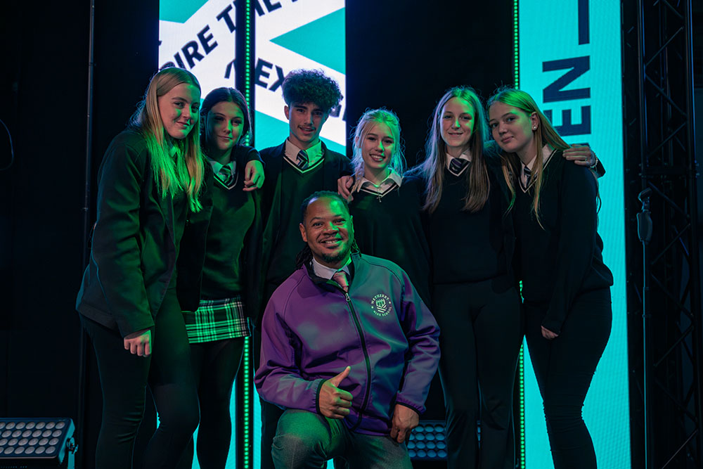 Six students in school uniform and a member of staff stood together smiling at the camera with Next Gen branding lit up behind the group