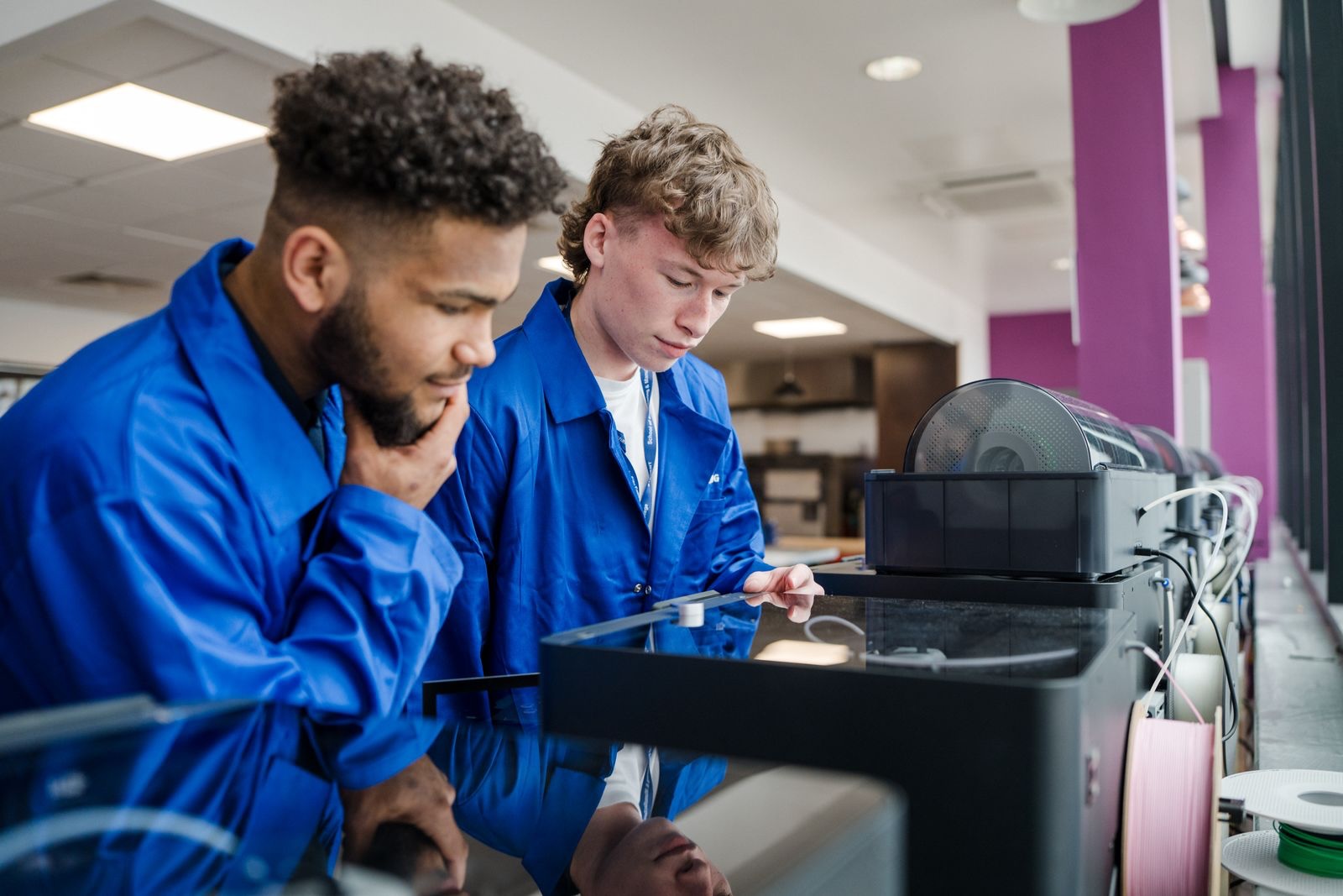 Two students wearing blue overalls working on an industrial 3d printer in the workshop