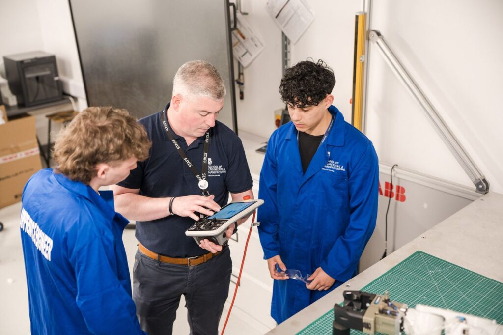Two students in their safety work gear stood listening to the instuctions and demonstration their tutor is giving them on how to work the machinery