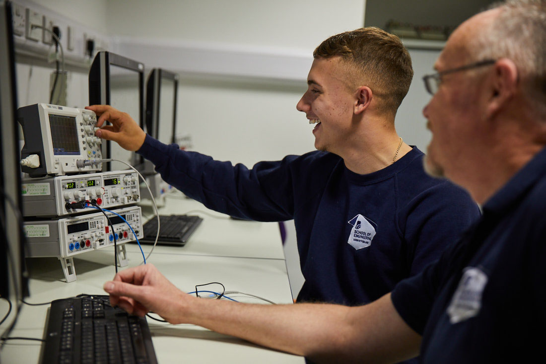 Leeds City College engineering student sat next to teacher, smiling whilst turning the dials on manufacturing equipment linked to computer