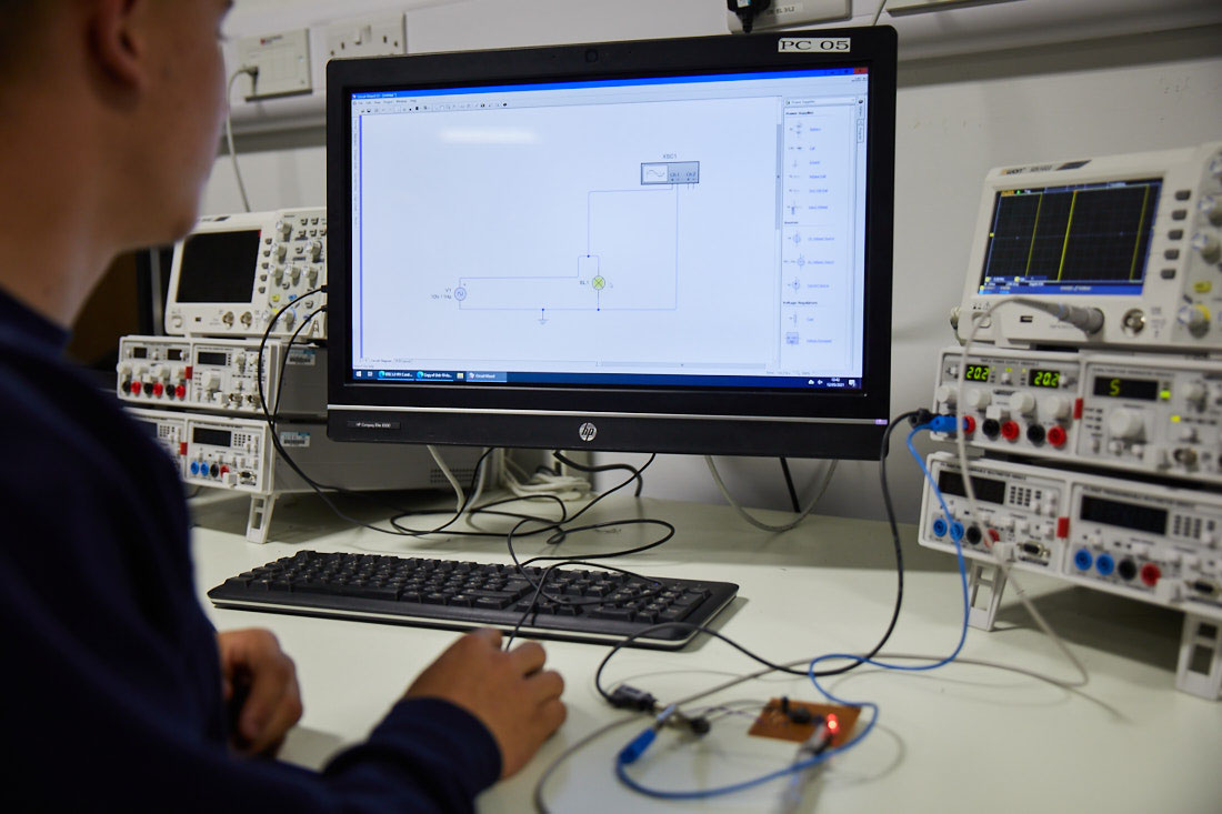 Student working on a design on desktop screen next to manufacturing equipment with lots of buttons and wires