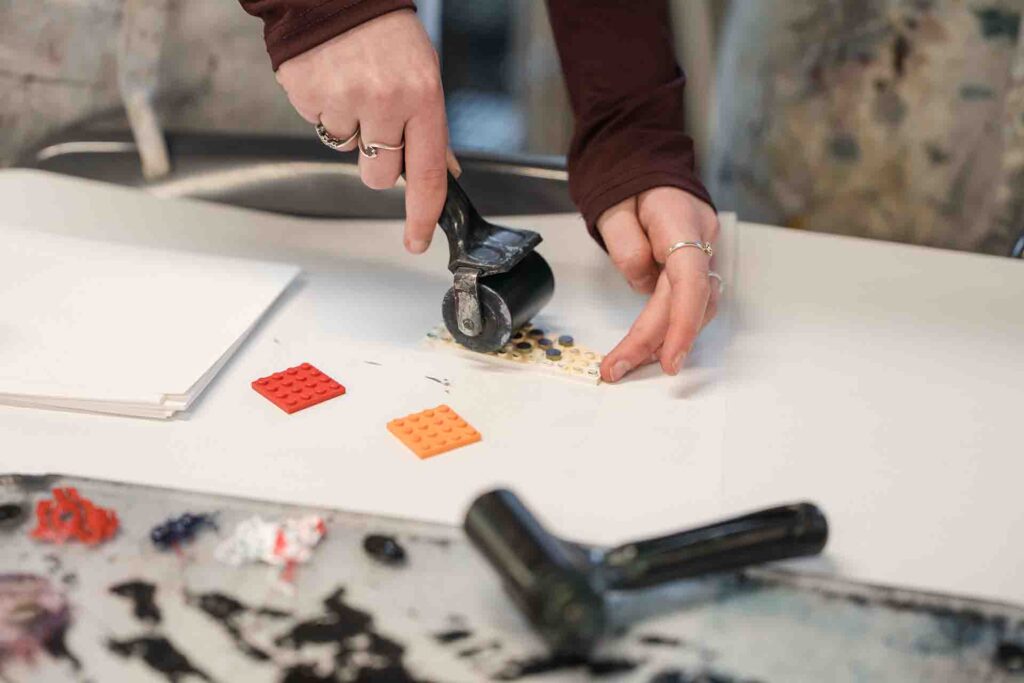 Close up of a person’s hands using a black brayer roller to apply ink over a plastic building block plate on a white table