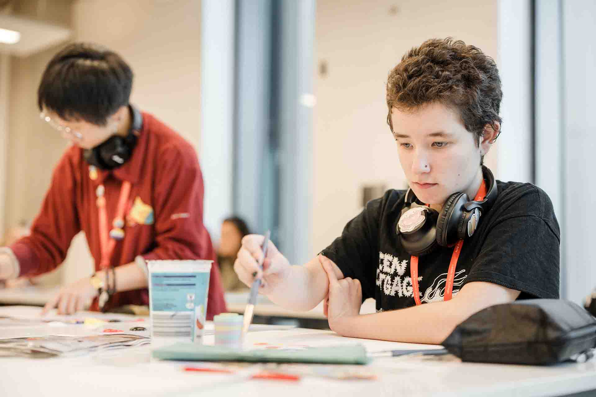 A focused student with short brown curly hair and headphones around their neck carefully paints at a white table. They are wearing a black graphic t-shirt and red lanyard. In the blurred background, another student in a red sweater works on their own art project