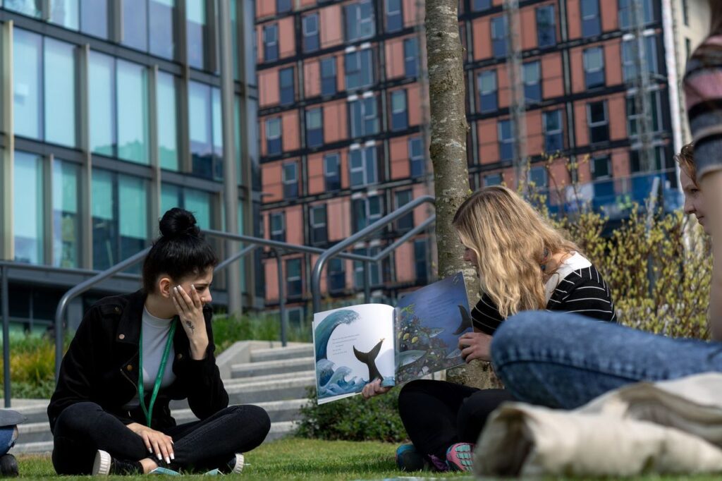 Students sat outside of Quarry Hill on the grass reading a children's book to the rest of the class