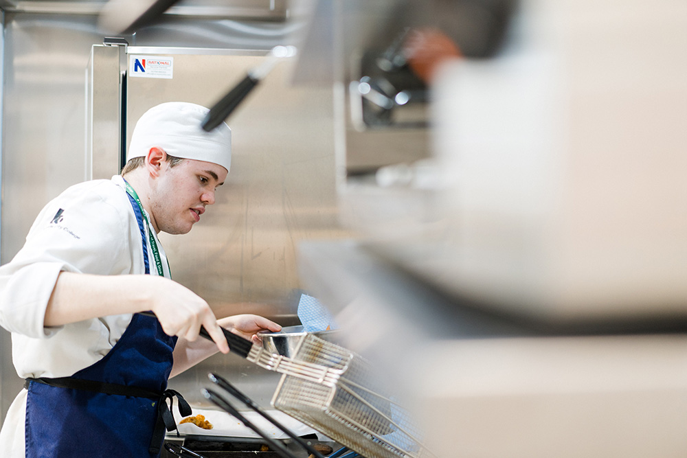 Student wearing chef overalls in Printworks kitchen moving chips out of cooker into a dish