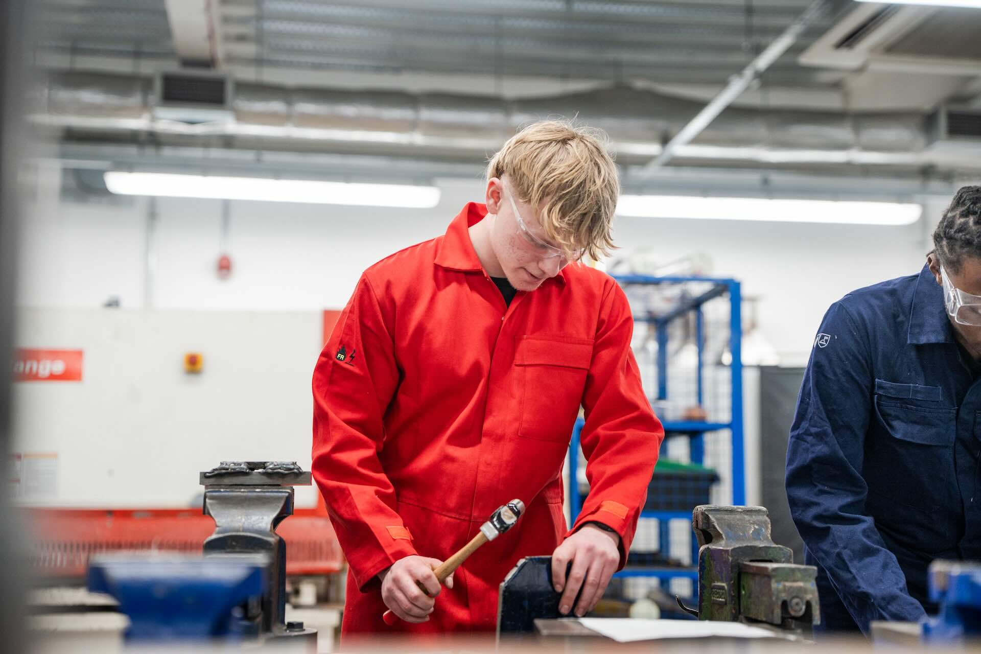 Student wearing bright red work overalls hammering a metal piece