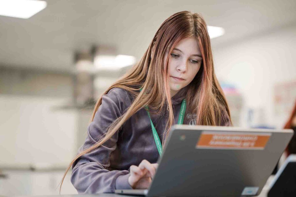 A young female student with long brown hair focused intently on typing on a grey laptop in a classroom setting