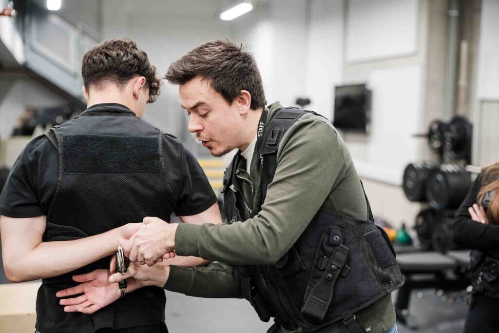 An instructor wearing a black tactical vest and a staff lanyard demonstrates how to apply handcuffs behind the back on a student in a black vest during a training exercise