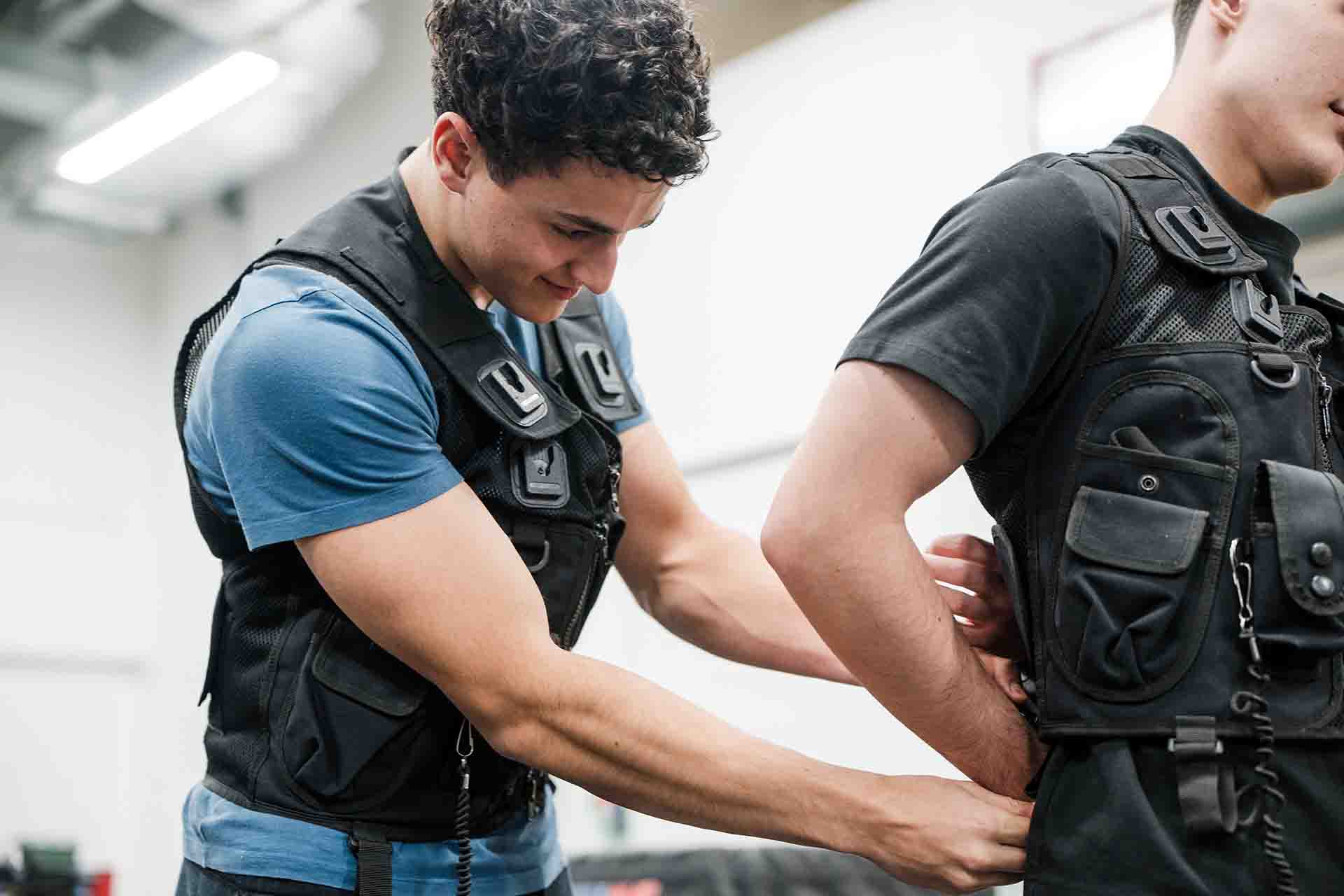 A male student wearing a blue shirt and tactical vest practices securing and adjusting a harness/ gear on another student's back during a public services training session