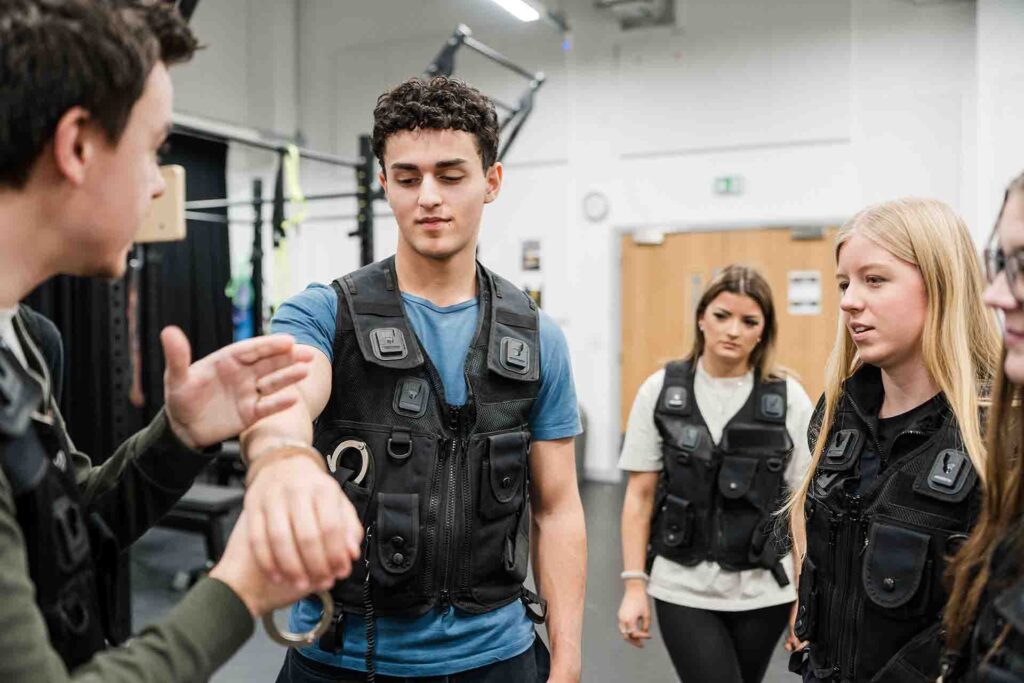 An instructor demonstrates handcuffing techniques on a male student wearing a tactical vest, while two female students in matching vests observe in a training room