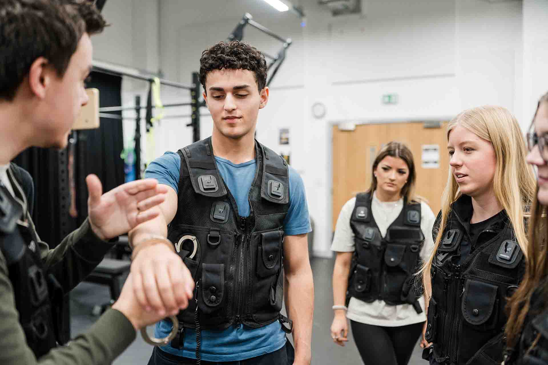 An instructor demonstrates handcuffing techniques on a male student wearing a tactical vest, while two female students in matching vests observe in a training room