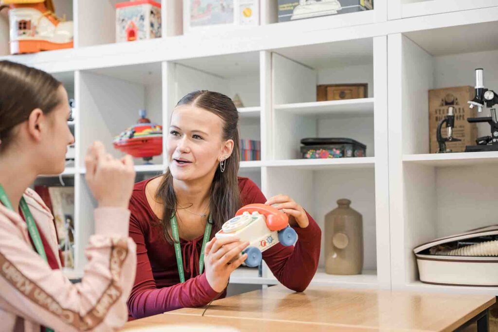 A female student wearing a red long-sleeved top holding a colourful toy telephone while talking with another student sitting across a table
