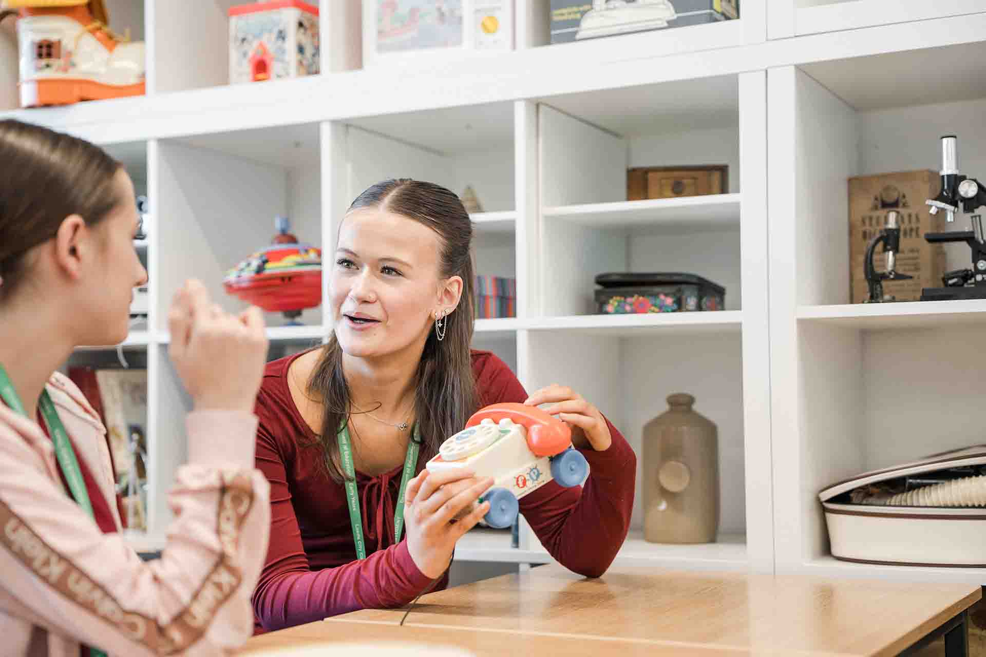 A female student wearing a red long-sleeved top holding a colourful toy telephone while talking with another student sitting across a table