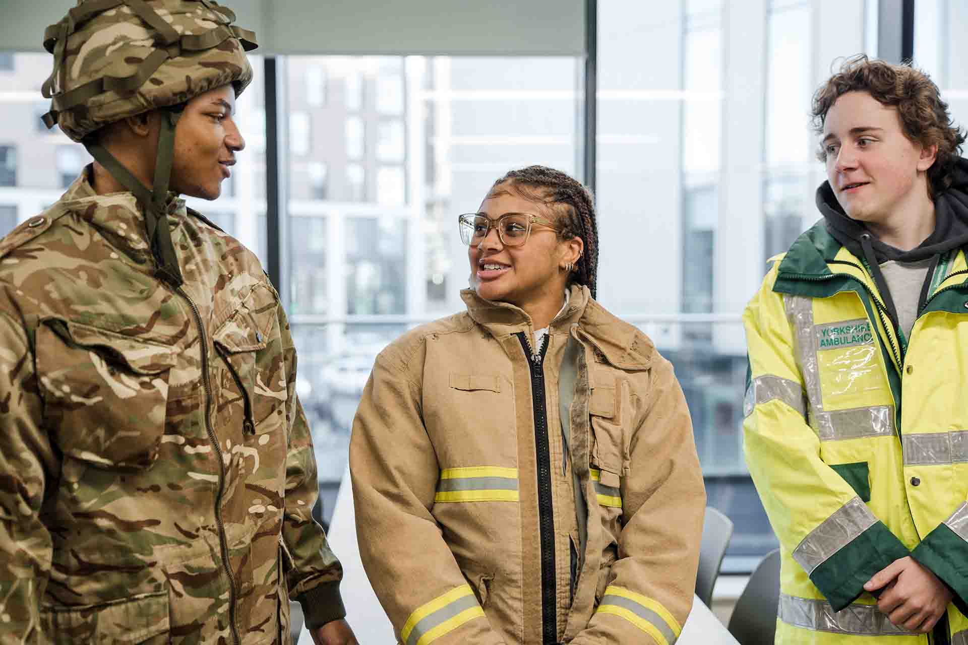 Three students wearing uniform jackets representing different emergency services, military camouflage, firefighter gear, and a high-visibility ambulance jacket, all smiling and chatting together