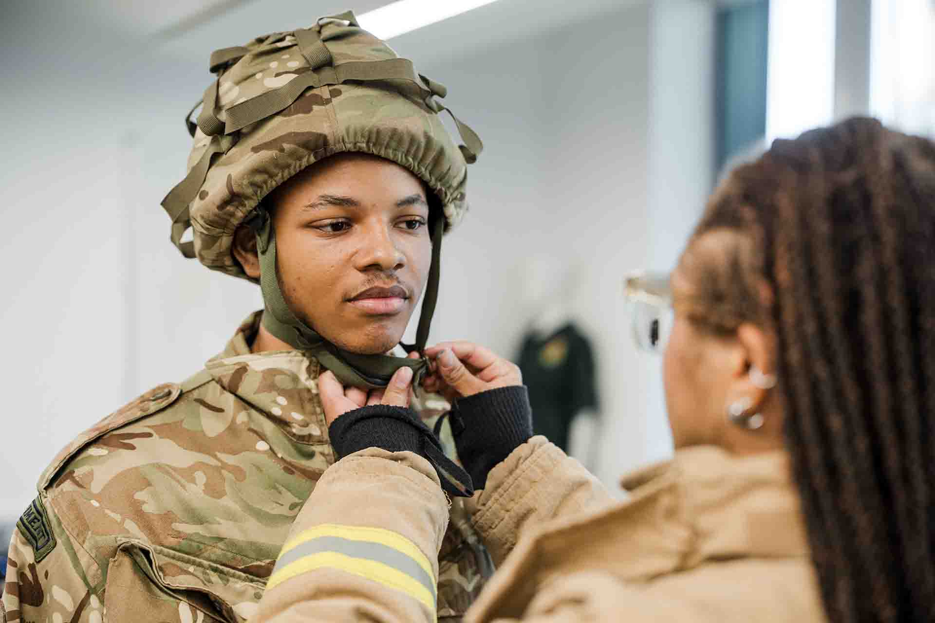 A student wearing a firefighter coat helps adjust the helmet chin strap for a fellow student wearing military camouflage gear
