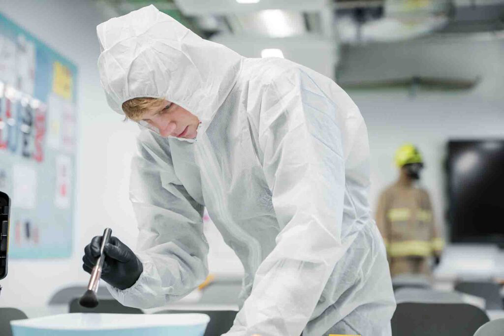 A student in a white protective forensics suit, hood, and black gloves carefully uses a makeup style brush to dusting for fingerprints and evidence on a surface