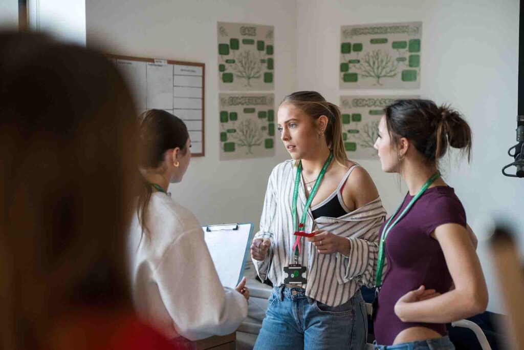 Three female healthcare students wearing lanyards standing in a medical training lab, engaging in a group discussion while one holds a clipboard