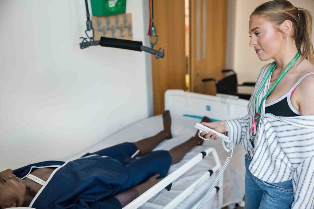 A female healthcare student standing beside a medical bed holds a white remote control to operate the patient hoist, while a training mannequin rests on the bed