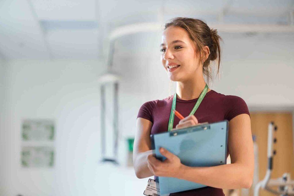 A smiling female student wearing a maroon top and green lanyard holding a blue clipboard and pen while looking slightly off camera in a training lab