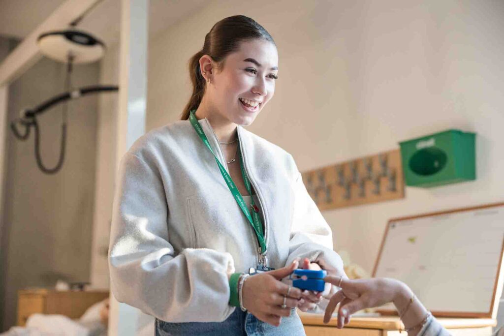 A smiling female student in a light grey jacket and green lanyard places a blue finger pulse oximeter onto another student's index finger in a medical training room